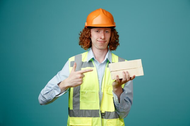 Joven constructor con uniforme de construcción y casco de seguridad sosteniendo dos ladrillos apuntándolos con el dedo índice sonriendo confiado de pie sobre fondo azul