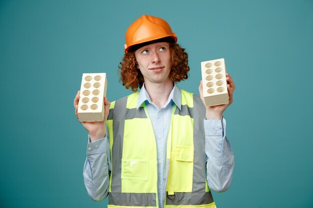 Joven constructor en uniforme de construcción y casco de seguridad sosteniendo dos ladrillos mirando hacia arriba con una sonrisa en la cara con expresión soñadora de pie sobre fondo azul.