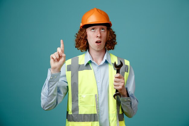 Joven constructor en uniforme de construcción y casco de seguridad sosteniendo una llave apuntando con el dedo índice hacia arriba mirando sorprendido de pie sobre fondo azul.