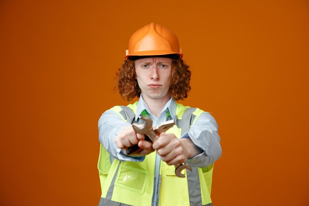 Joven constructor en uniforme de construcción y casco de seguridad sosteniendo llaves cruzando las manos mirando a la cámara con el ceño fruncido haciendo un gesto de parada de pie sobre fondo naranja