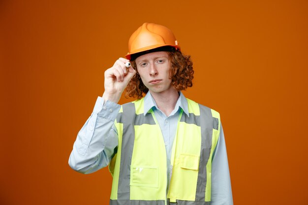 Joven constructor en uniforme de construcción y casco de seguridad sosteniendo marcador mirando a la cámara con cara seria sosteniendo de pie sobre fondo naranja