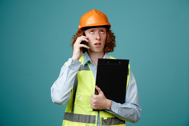 Joven constructor en uniforme de construcción y casco de seguridad sosteniendo portapapeles hablando por teléfono móvil mirando confundido de pie sobre fondo azul