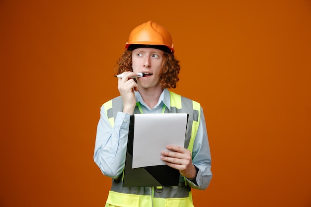 Joven constructor en uniforme de construcción y casco de seguridad sosteniendo portapapeles y marcador mirando a un lado con expresión pensativa pensando de pie sobre fondo naranja