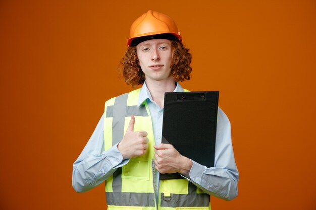 Joven constructor en uniforme de construcción y casco de seguridad sosteniendo portapapeles mirando a la cámara sonriendo confiado mostrando el pulgar hacia arriba de pie sobre fondo naranja