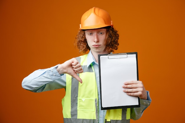 Joven constructor en uniforme de construcción y casco de seguridad sosteniendo portapapeles con páginas en blanco que parece disgustado mostrando el pulgar hacia abajo de pie sobre fondo naranja