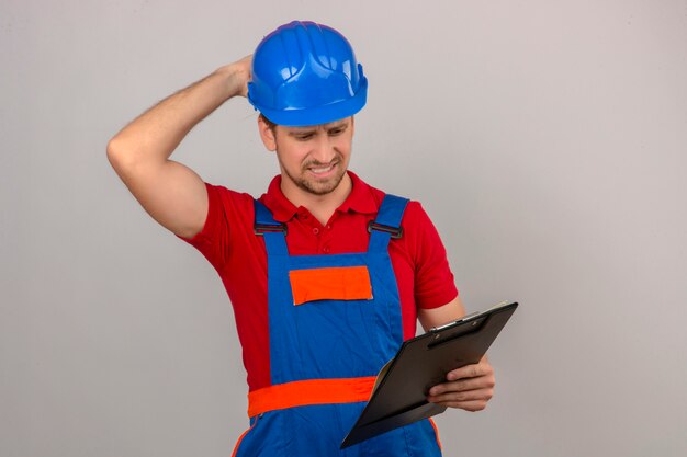 Joven constructor en uniforme de construcción y casco de seguridad sosteniendo portapapeles pensando con la mano en la cabeza mirada confundida teniendo dudas sobre la pared blanca aislada