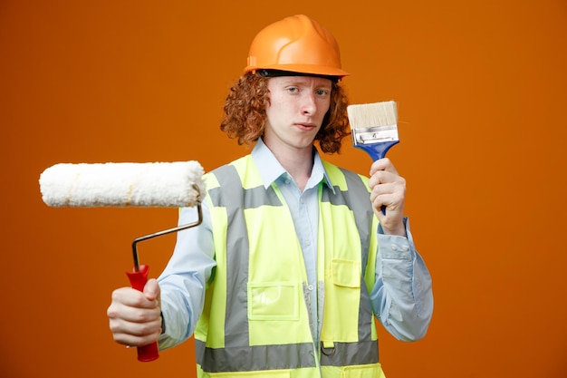 Joven constructor en uniforme de construcción y casco de seguridad sosteniendo rodillo de pintura y cepillo mirando a la cámara con cara seria de pie sobre fondo naranja