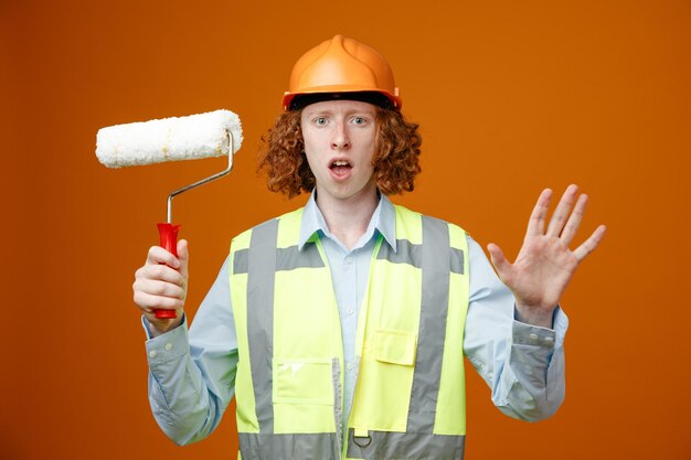 Joven constructor en uniforme de construcción y casco de seguridad sosteniendo un rodillo de pintura mirando a la cámara sorprendido levantando el brazo preocupado de pie sobre fondo naranja