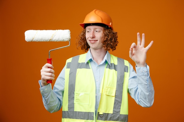 Joven constructor en uniforme de construcción y casco de seguridad sosteniendo un rodillo de pintura mirándolo con una sonrisa en la cara mostrando el signo de ok de pie sobre fondo naranja