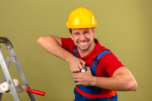 Joven constructor en uniforme de construcción y casco de seguridad tratando de abrir la pintura puede ser difícil de abrir el concepto sobre la pared verde aislada