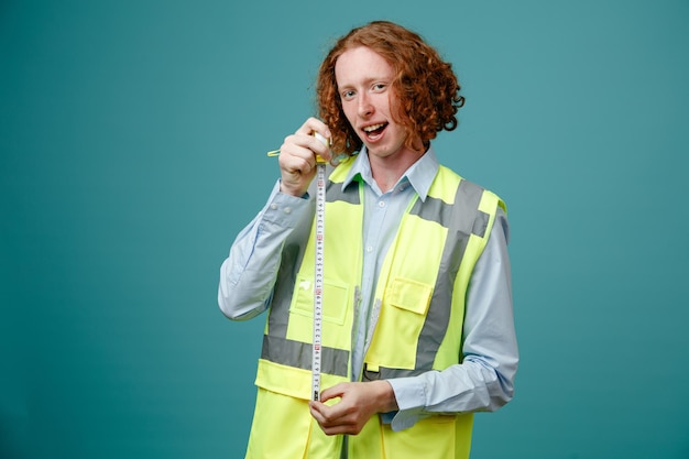 Foto gratuita joven constructor en uniforme de construcción sosteniendo cinta métrica mirando a cámara feliz y positiva sonriendo ampliamente de pie sobre fondo azul.