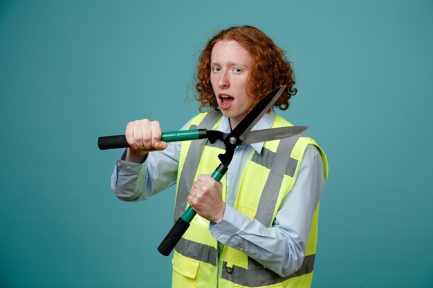 Joven constructor en uniforme de construcción sosteniendo tijeras mirando a la cámara emocionado y feliz de pie sobre fondo azul.