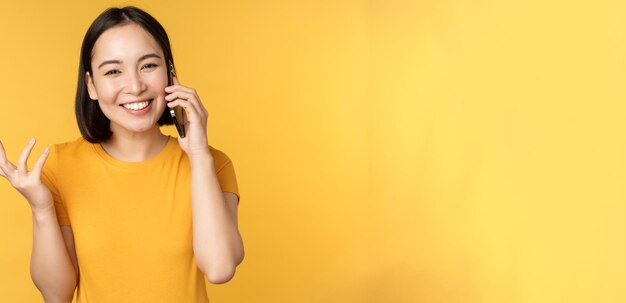 Joven coreana hablando por teléfono móvil mujer asiática llamando a un teléfono inteligente de pie sobre bac amarillo