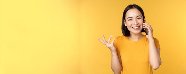 Joven coreana hablando por teléfono móvil mujer asiática llamando a un teléfono inteligente de pie sobre bac amarillo