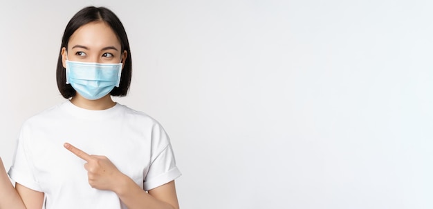 Joven coreana con mascarilla médica apuntando con el dedo a la izquierda y mirando el logotipo que muestra publicidad o pancarta de pie sobre fondo blanco