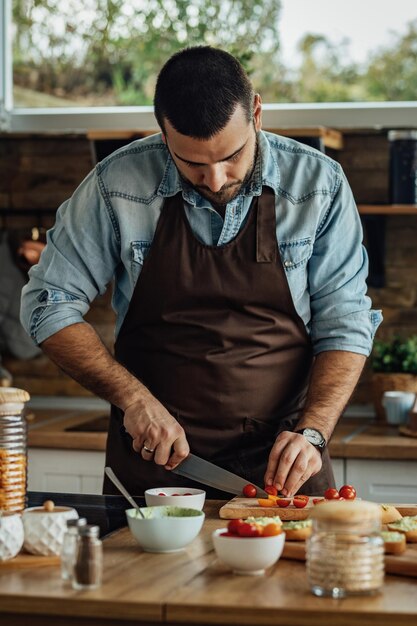 Joven cortando tomate cherry en la tabla de cortar mientras prepara comida saludable en la cocina.