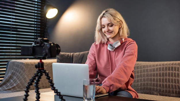 Joven creador de contenido sonriente chica rubia con auriculares trabajando en su computadora portátil sobre la mesa con cámara