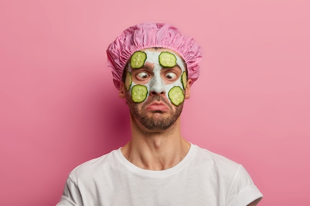 Joven cruza los ojos, aplica mascarilla de crema en la cara con pepino, tiene sesión de belleza, usa gorro de baño