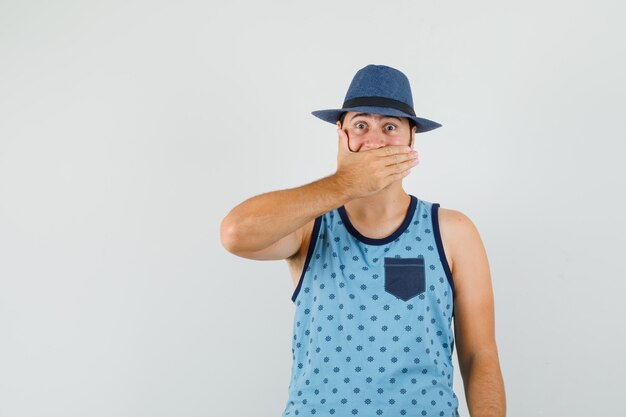 Joven cubriendo la boca con la mano en camiseta azul, sombrero y mirando consternado,