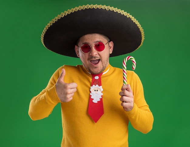 Foto gratuita joven de cuello alto amarillo y gafas con divertida corbata roja con sombrero negro sosteniendo bastón de caramelo con cara feliz sonriendo mostrando los pulgares para arriba de pie sobre la pared verde