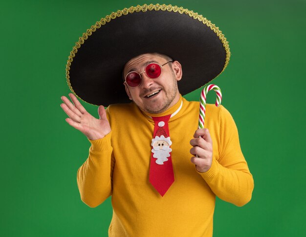 Foto gratuita joven de cuello alto amarillo y gafas con divertida corbata roja con sombrero negro sosteniendo bastón de caramelo con cara feliz sonriendo de pie sobre la pared verde