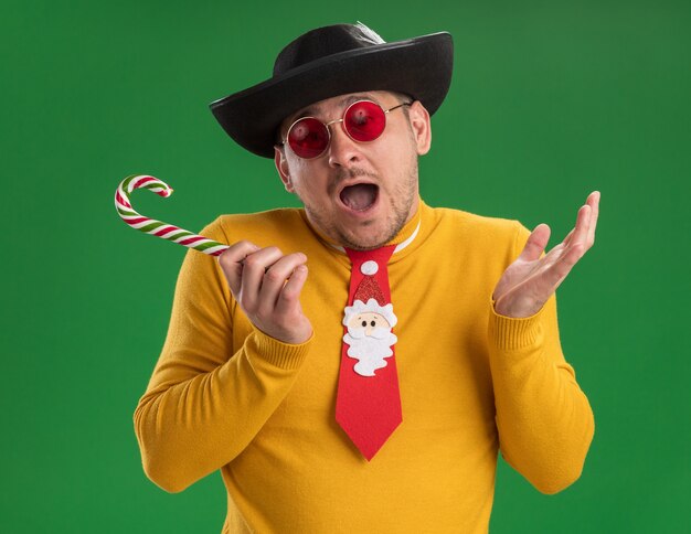 Joven de cuello alto amarillo y gafas con divertida corbata roja con sombrero negro sosteniendo bastón de caramelo feliz y emocionado de pie sobre la pared verde