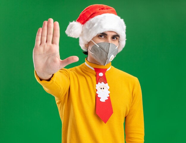 Joven de cuello alto amarillo y gorro de Papá Noel con corbata divertida con máscara protectora facial mirando a la cámara con cara seria haciendo gesto de parada con la mano abierta de pie sobre fondo verde