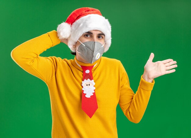 Joven de cuello alto amarillo y gorro de Papá Noel con corbata divertida con máscara protectora facial mirando a la cámara sorprendido con el brazo levantado sobre fondo verde