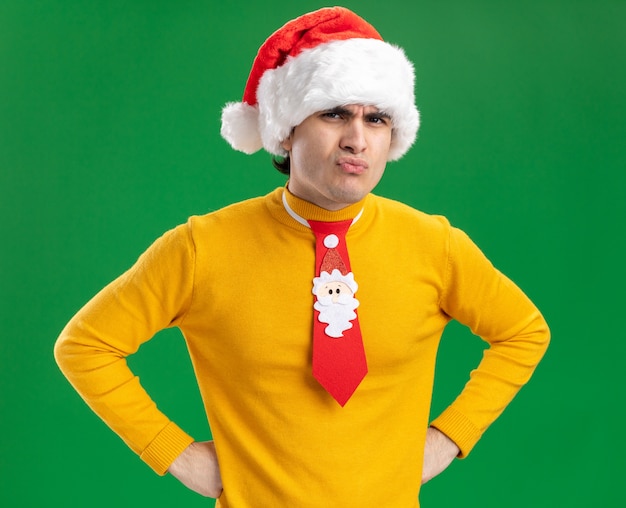Joven de cuello alto amarillo y gorro de Papá Noel con corbata divertida mirando a cámara disgustado con cara enojada con las manos en la cadera de pie sobre fondo verde