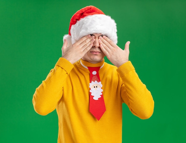 Joven de cuello alto amarillo y gorro de Papá Noel con corbata divertida que cubre los ojos con las manos de pie sobre la pared verde