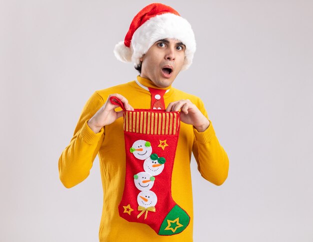 Joven de cuello alto amarillo y gorro de Papá Noel con corbata divertida sosteniendo calcetín navideño mirando a cámara asombrado y sorprendido de pie sobre fondo blanco.