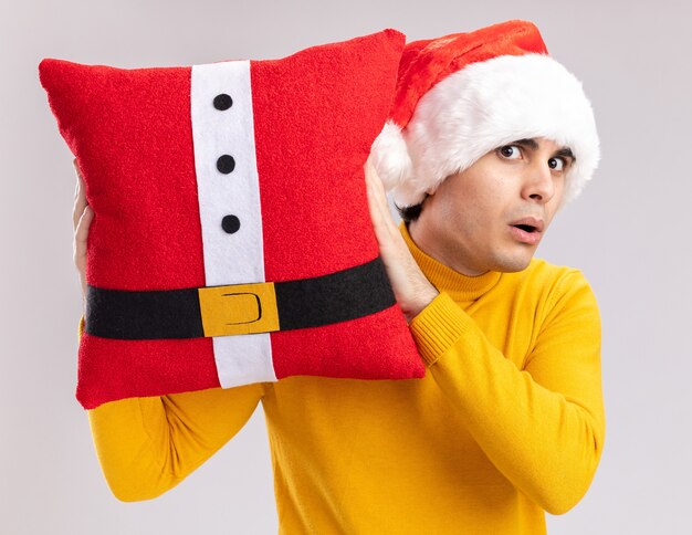 Joven de cuello alto amarillo y gorro de Papá Noel sosteniendo la almohada de Navidad mirando a la cámara sorprendido de pie sobre fondo blanco.