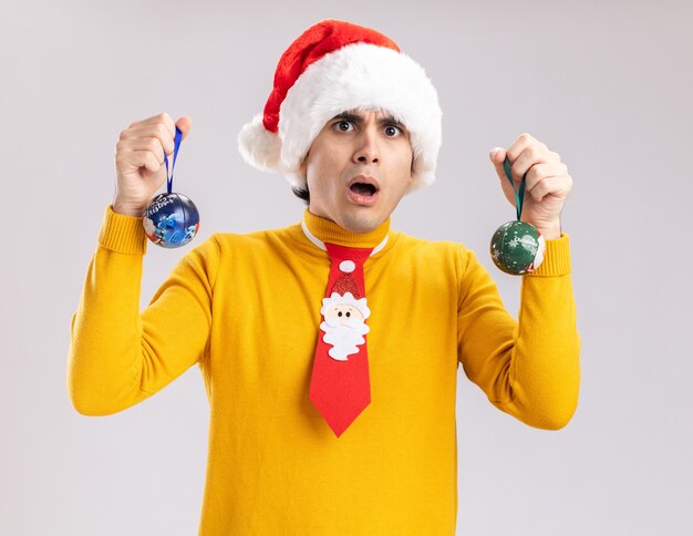 Joven de cuello alto amarillo y gorro de Papá Noel sosteniendo bolas de navidad asombrado y sorprendido de pie sobre la pared blanca