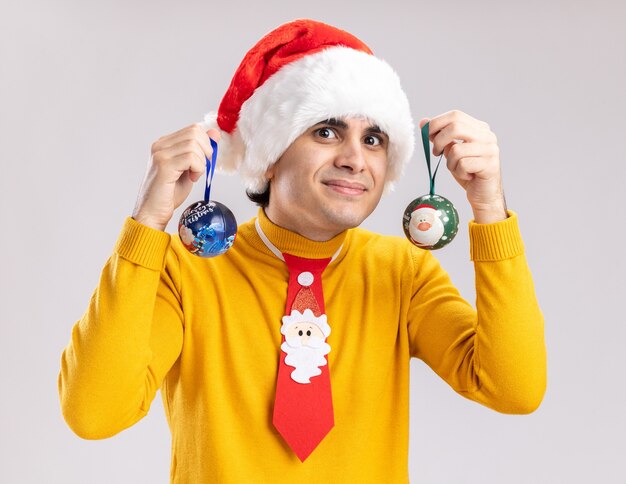 Joven de cuello alto amarillo y gorro de Papá Noel sosteniendo bolas de navidad feliz y sonriente de pie sobre la pared blanca