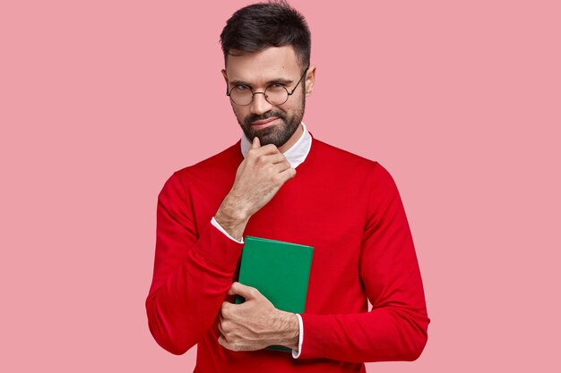 Un joven cunny tiene la intención de hacer algo, sostiene la barbilla, lleva un cuaderno verde para escribir notas, usa un suéter rojo, gafas ópticas