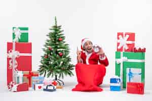 Foto gratuita joven curioso celebrar año nuevo o vacaciones de navidad sentado en el suelo y sosteniendo el reloj cerca de regalos y árbol de navidad decorado