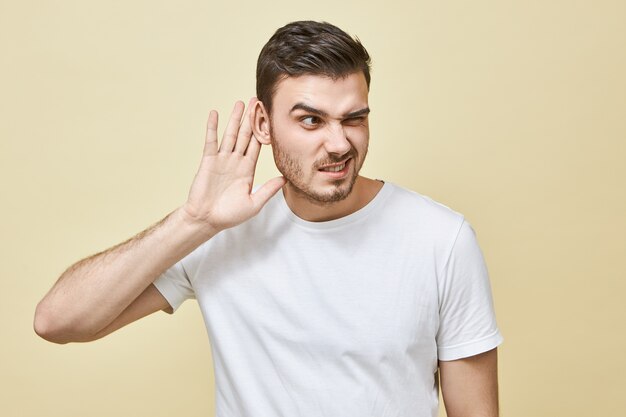 Joven curioso con cerdas manteniendo la mano en la oreja mientras escucha a escondidas, escucha el secreto, tiene una expresión facial concentrada, tratando de escuchar todo. Concepto de chismes y rumores
