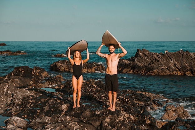 Joven dama y chico con tablas de surf en las cabezas de roca cerca del mar