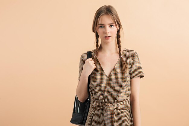 Joven dama hermosa con dos trenzas en vestido de tweed sosteniendo una mochila negra en el hombro mientras mira soñadoramente a la cámara sobre un fondo beige