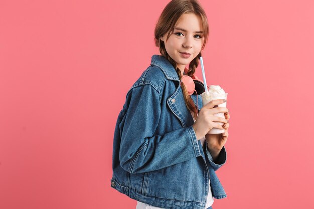 Joven dama muy sonriente con dos trenzas en una chaqueta de mezclilla con auriculares sosteniendo un batido en las manos mientras mira felizmente a la cámara sobre un fondo rosa