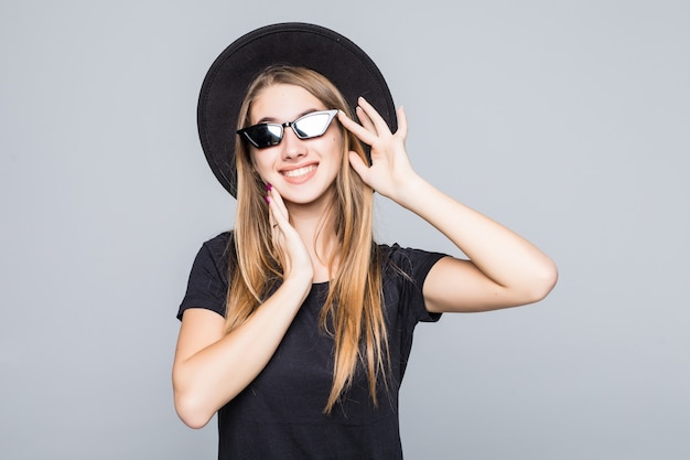 Joven dama muy sonriente feliz con gafas de sol brillantes vestida con sombrero negro, camiseta negra y pantalón oscuro aislado sobre fondo gris