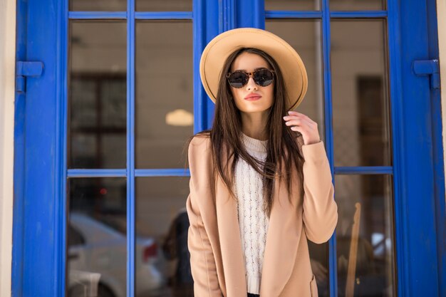 Joven dama vestida casual con sombrero brillante y gafas de sol está de pie delante de la puerta azul