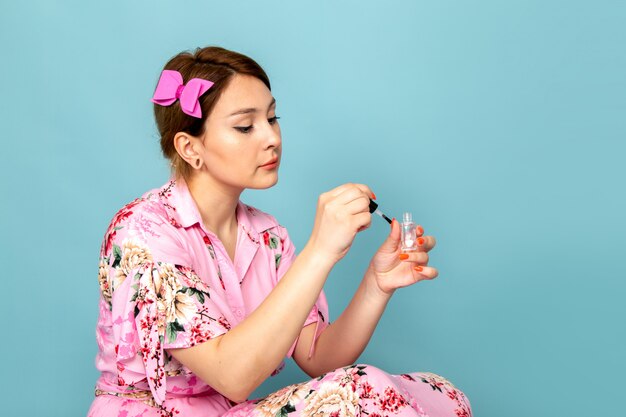 Una joven dama de vista frontal en vestido rosa diseñado con flores pintando sus uñas en azul