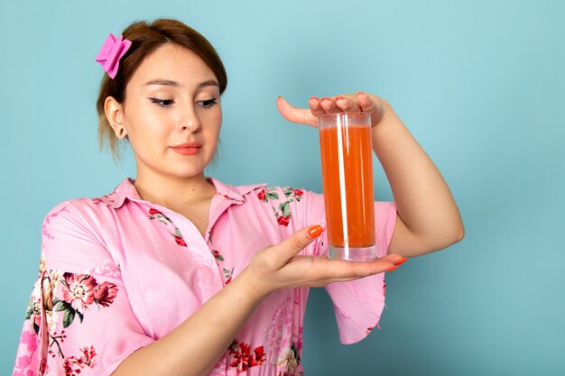 Una joven dama de vista frontal en vestido rosa diseñado con flores sosteniendo agua de píldora de color naranja sobre azul