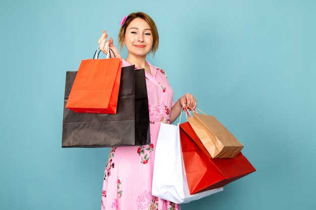 Una joven dama de vista frontal en vestido rosa diseñado con flores sosteniendo paquetes de compras y sonriendo en azul