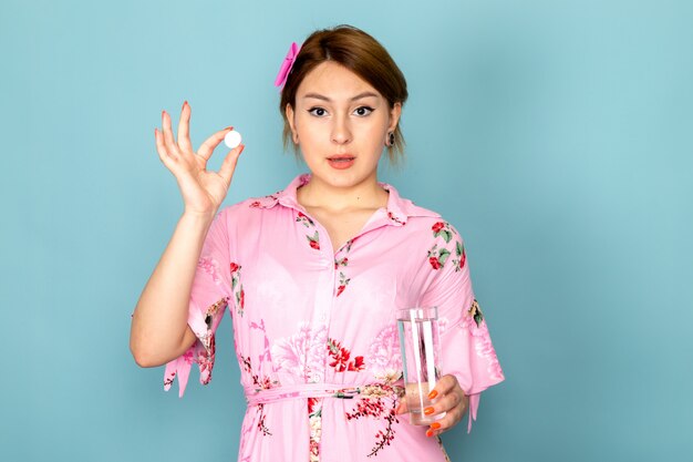 Una joven dama de vista frontal en vestido rosa diseñado con flores sosteniendo la píldora y un vaso de agua en azul