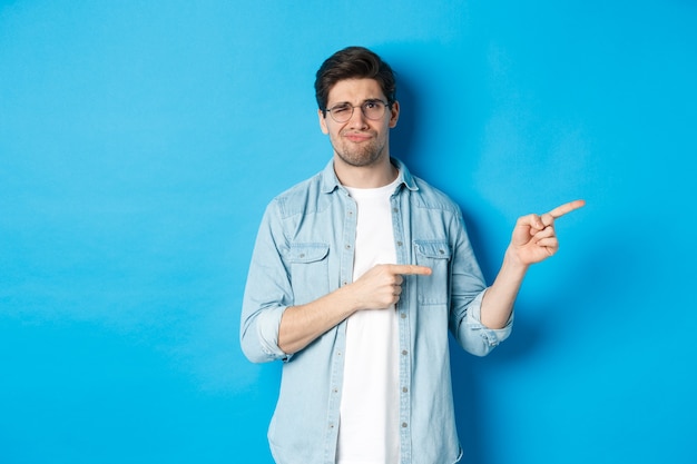 Joven decepcionado con gafas apuntando con el dedo a la derecha en el espacio de la copia, mostrando pancarta promocional y sonriendo insatisfecho, juzgando mal producto, de pie sobre fondo azul