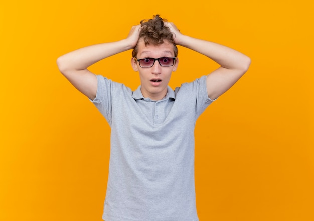 Joven decepcionado de gafas negras con camisa de polo gris tocando su cabeza confundida sobre naranja