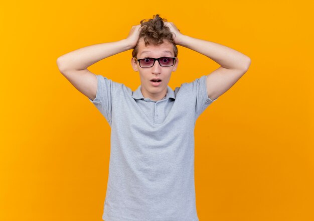 Joven decepcionado de gafas negras con camisa de polo gris tocando su cabeza confundida sobre naranja