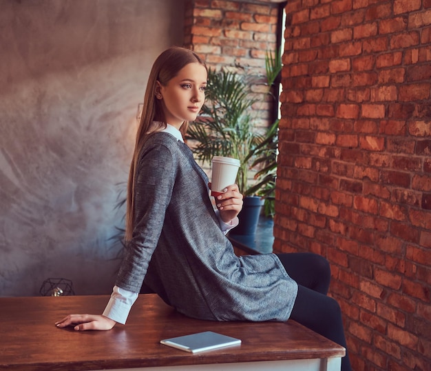 Una joven delgada y sexy con un vestido gris sostiene una taza de café sentada